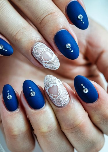 Elegant blue matte nails with intricate white lace designs and sparkling rhinestones.