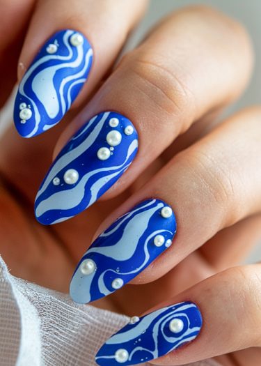 Vibrant cobalt blue nail art features white wavy designs and elegant pearl embellishments.