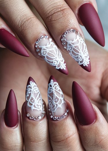 Elegant burgundy matte stiletto nails with intricate lace design and sparkling rhinestones.