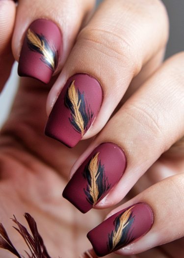 Elegant burgundy matte nail art with gold and black feather designs for stylish manicures.