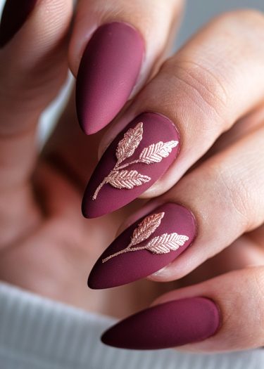 Elegant burgundy matte stiletto nails with gold leaf design, perfect for modern nail art.