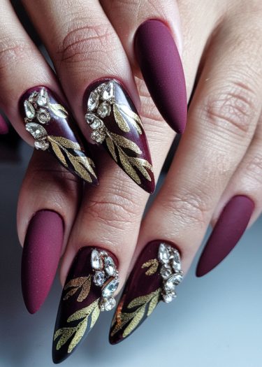 Elegant burgundy almond nails with gold leaf designs and rhinestones for a luxurious manicure.