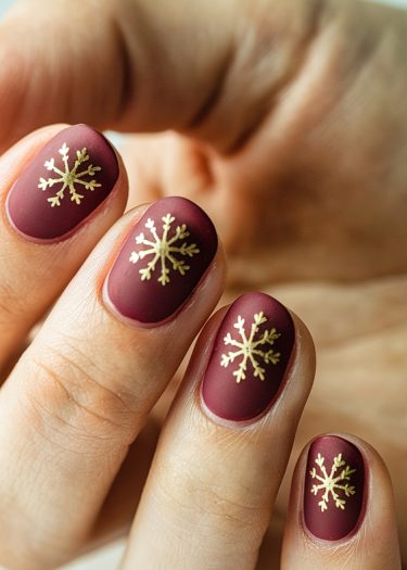 Elegant burgundy nails adorned with golden snowflake designs, perfect for winter and holiday celebrations.