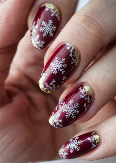 Elegant burgundy nails with intricate white snowflakes and gold accents for a festive winter look.