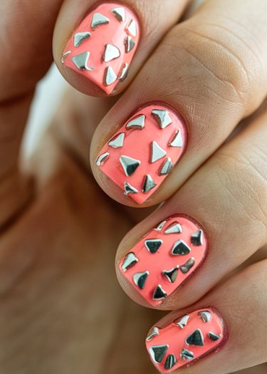 Vibrant coral pink nail art with metallic silver triangle embellishments for a chic design.