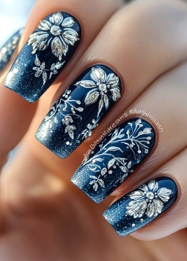 Elegant navy blue ombre nails with intricate silver floral designs and shimmering glitter accents.