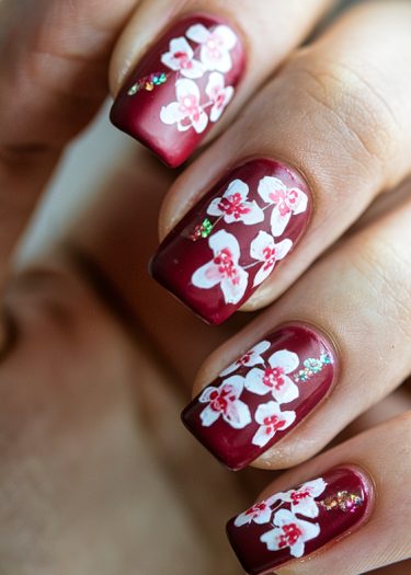 Elegant burgundy nail art featuring intricate white floral designs and sparkling green accents.