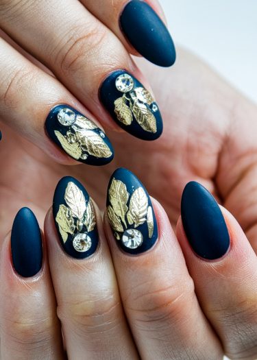 Elegant deep blue almond nails with gold leaf designs and sparkling rhinestones for a luxurious look.