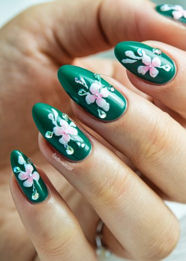 Elegant deep green almond-shaped nails adorned with pink floral designs and sparkling rhinestones.