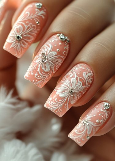 Elegant peachy-pink nails with intricate white floral designs and sparkling rhinestones for a stunning manicure.