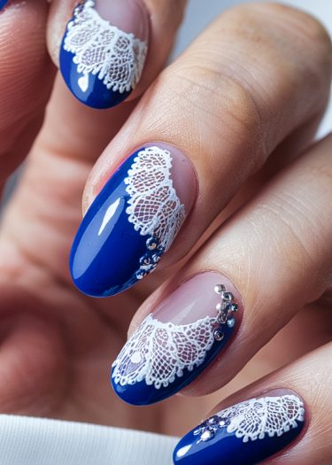 Elegant royal blue nail design with intricate white lace patterns and sparkling rhinestones.