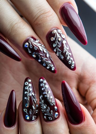 Elegant burgundy stiletto nails with intricate silver nail art and sparkling rhinestones.