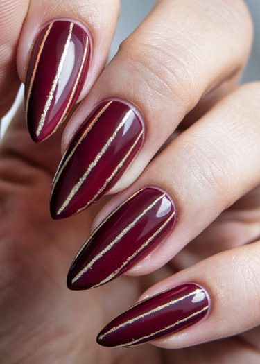 Elegant burgundy manicure with gold accents, showcasing long almond-shaped nails in a glossy finish.