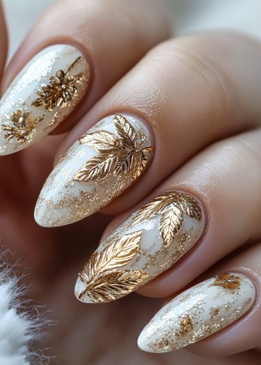 Elegant gold leaf nail art on almond-shaped, creamy white nails with floral designs.