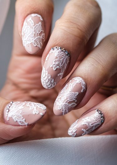 Elegant nude pink nail art featuring intricate white lace designs and sparkling rhinestones.