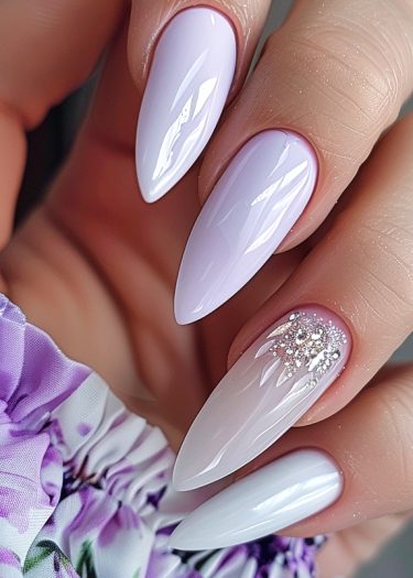 Elegant lilac almond nails with rhinestone accents and floral fabric backdrop.