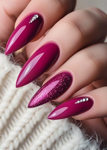 Elegant stiletto magenta nails with rhinestones and glitter on a cozy fabric background.