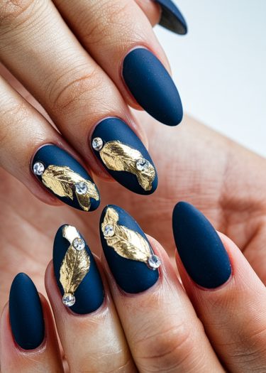 Elegant matte navy nails adorned with gold foil accents and rhinestones for a sophisticated look.