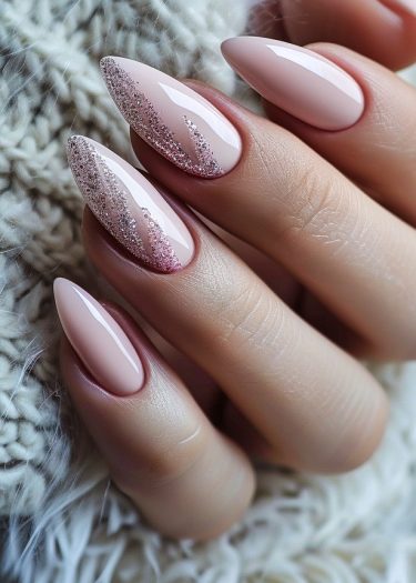 Elegant almond-shaped nude pink nails with glitter accents on a soft fabric background.