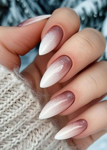 Elegant almond-shaped ombré nails with glitter against a wintery pine tree background.