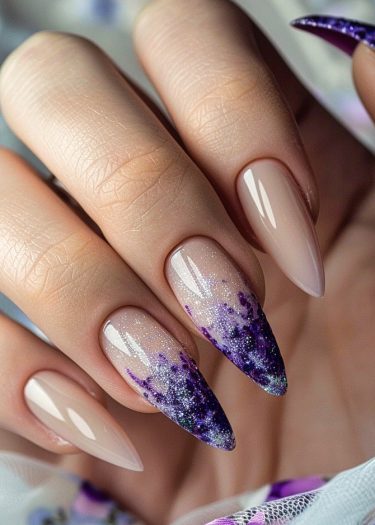 Elegant stiletto nails feature a purple ombre design with glitter on a manicured hand.