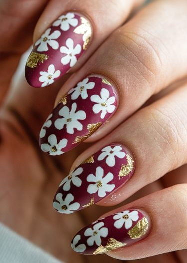 Elegant burgundy almond nails with white floral designs and golden accents for a sophisticated look.