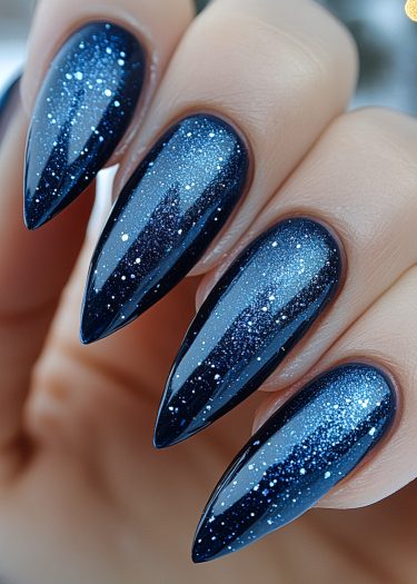 Elegant stiletto nails with galaxy-inspired dark blue lacquer and sparkling glitter design.