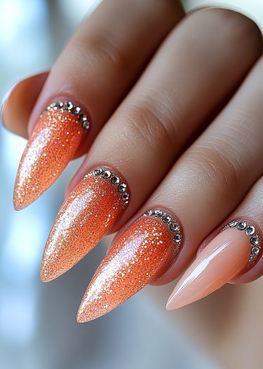 Elegant stiletto nails adorned with vibrant orange glitter and sparkling rhinestones.