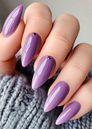 Elegant glossy violet almond nails with embellishments resting on a cozy gray knitted fabric.