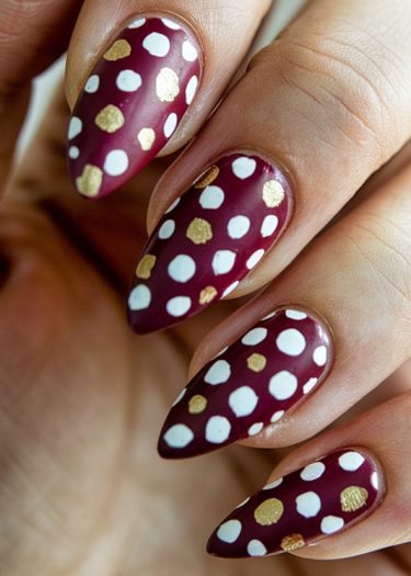 Elegant burgundy stiletto nails with playful white polka dots and shimmering gold accents.