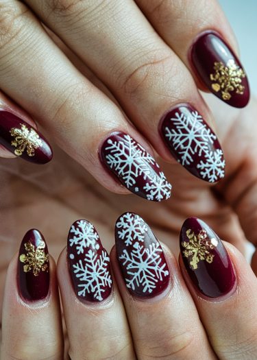 Elegant burgundy nail art with intricate white snowflakes and gold accents for winter.