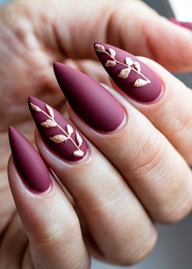 Elegant maroon stiletto nails with gold leaf design showcasing sophisticated nail art.