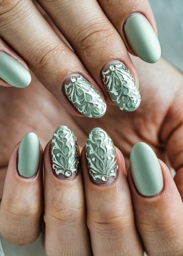 Elegant sage green matte and glossy almond nails with embossed floral designs and rhinestone accents.