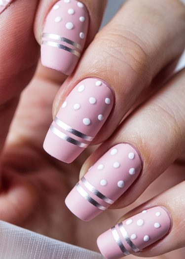 Elegant matte pink nails with white polka dots and silver stripes for a chic look.