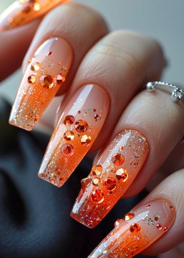 Elegant orange gradient nail art with rhinestones and glitter for a glamorous look.