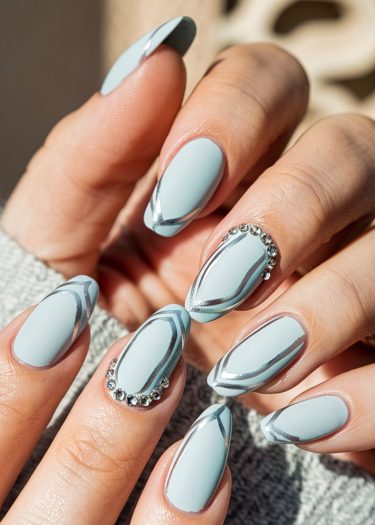 Elegant pastel blue almond nails with intricate silver designs and sparkling rhinestones.