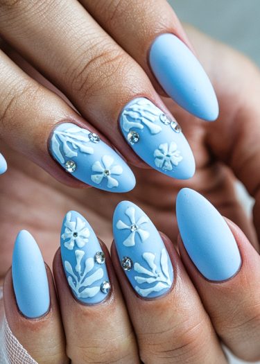 Elegant pastel blue matte nails with intricate white 3D designs and sparkling rhinestones.