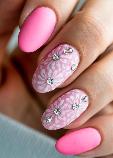 Vibrant pink almond nails with floral designs and rhinestones showcase modern nail art trends.