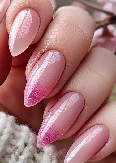 Elegant almond-shaped pastel pink nails with glitter gradient, featuring intricate white designs.