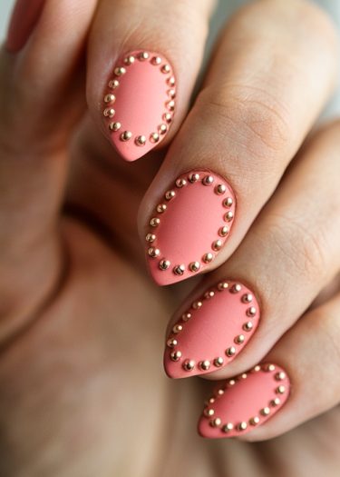 Elegant coral pink almond nails with gold studs for a luxurious manicure look.