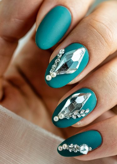 Elegant matte teal stiletto nails adorned with silver gemstones, pearls, and intricate designs.