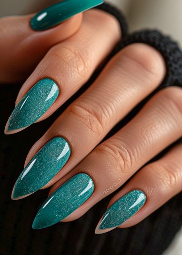 Beautifully manicured teal stiletto nails with shimmer on a smooth hand against dark fabric.