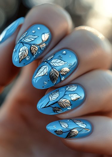 Elegant stiletto nails with vibrant blue polish and metallic leaf designs adorned with rhinestones.