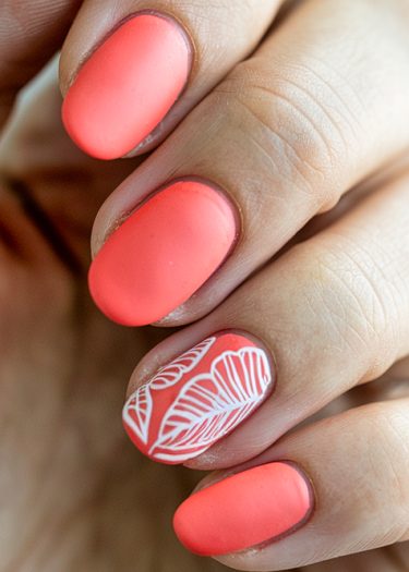 Close-up of vibrant coral pink matte nails with intricate white leaf design on ring finger.