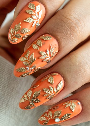 Vibrant orange stiletto nails with gold leaf designs and rhinestones for elegant nail art.