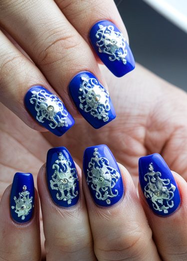 Elegant royal blue artificial nails adorned with intricate silver designs and sparkling gems.